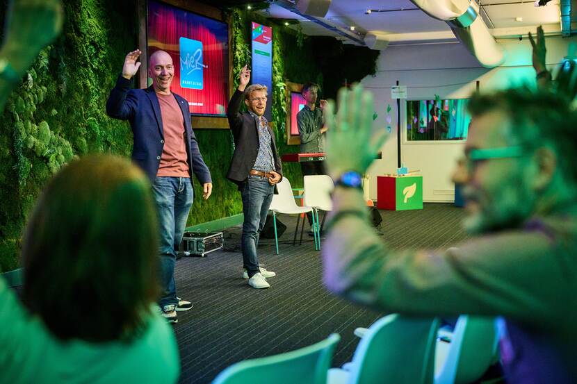 Twee sprekers staan met opgestoken handen voor de zaal, ook mensen uit het publiek steken hun hand op